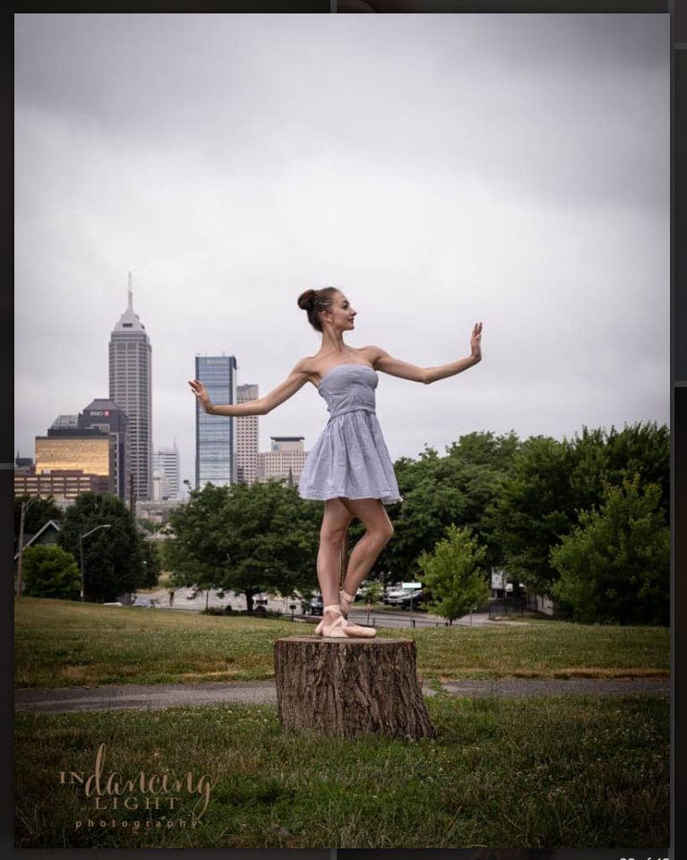 A ballerina poses on a tree stump in a park with the Indianapolis skyline in the background.