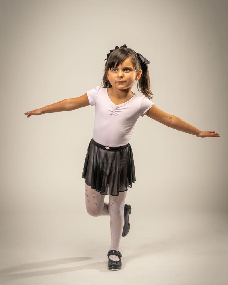 Portrait of an Indianapolis child dancing with outstretched arms and standing on one foot wearing a black dance skirt.