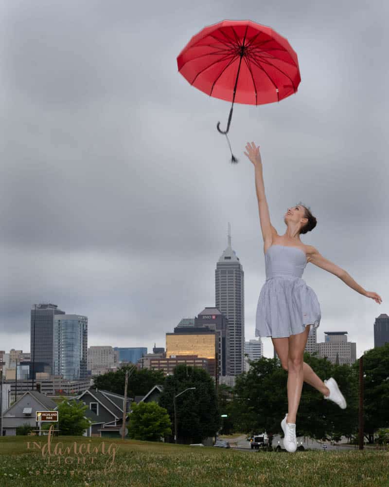 Ballerina leaps into the air to catch a red umbrella. The Indianapolis skyline is in the background