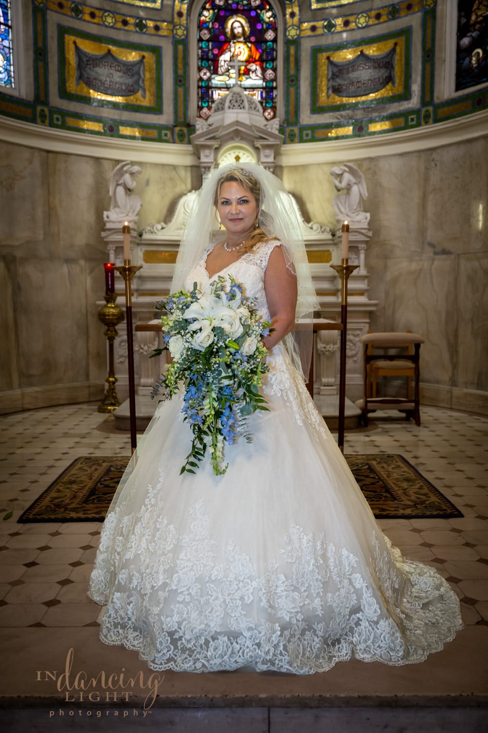 Bride stands facing at the front of the chapel following her wedding in the Indianapolis Cathedral. The alter and stained glass windows are in the background.