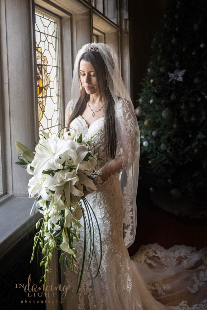 Bride in a beautiful gown holds her bouquet of white lilies as she stands by a window in Indianapolis Laurel Hall.