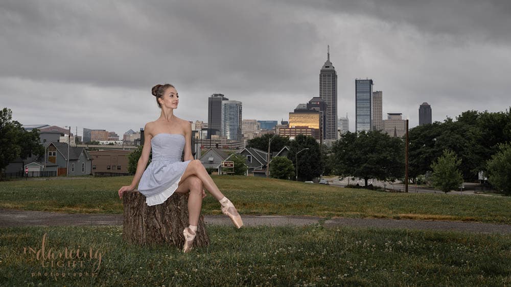 Ballerina rests on a tree stump with the Indianapolis skyline in the background.