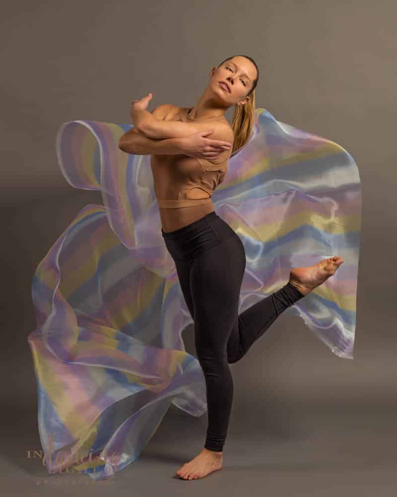 Dancer stands on one leg with rainbow colored fabric behind her