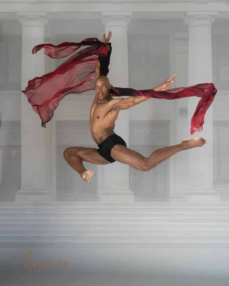 male dancer leaps with a flowing red fabric wrapped around one arm