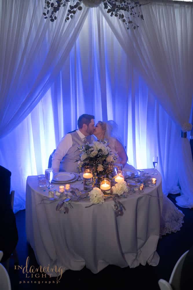 Bride and groom kiss as they are seated at their wedding reception table. Candles and bouquet are on the table. Curtains lit with blue light are in the background.